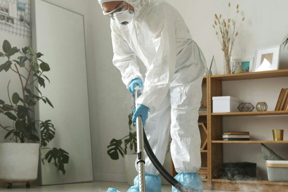 Person in protective gear vacuuming a room with plants and shelves in the background.